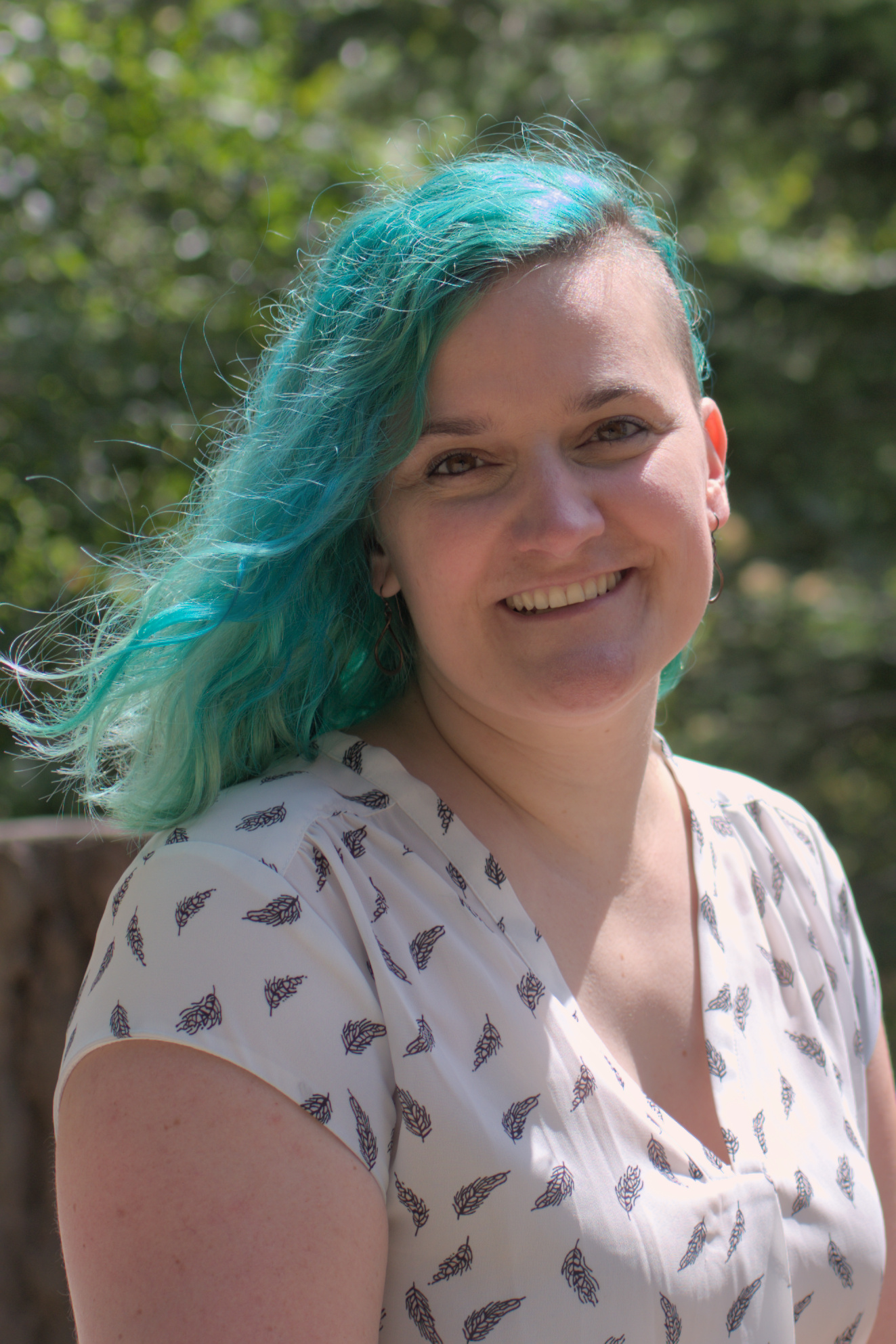 A white woman with teal hair wearing a white shirt smiling at the camera.